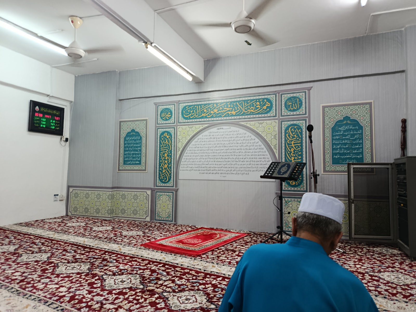 Vista Angkasa: A prayer room (surau) with Islamic calligraphy on the wall, a prayer mat, Quran stand and microphone, with a man in a blue robe and white cap kneeling