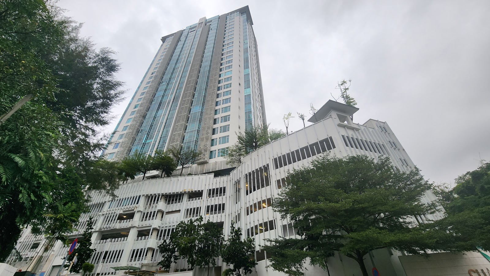 Suasana Bangsar: A tall high-rise residential tower viewed from ground level, surrounded by trees, with cloudy sky in the background
