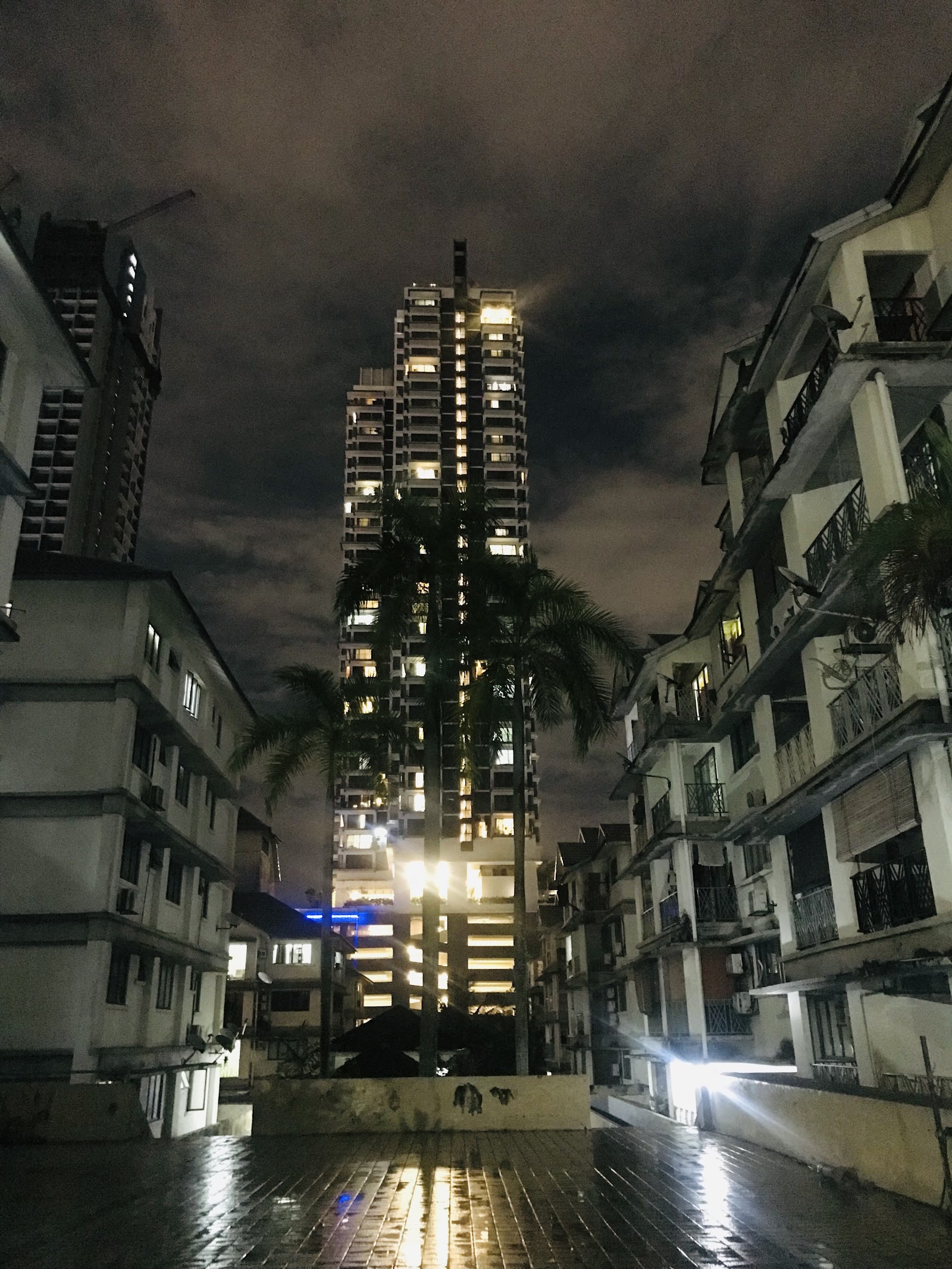 Bangsar Permai: Night view of a tall residential tower surrounded by palm trees, flanked by lower apartment buildings, taken from a courtyard