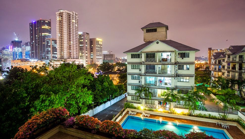 Bangsar Permai: Night view of a residential building with pool and city skyline in background, taken from an elevated balcony with flowering plants in foreground