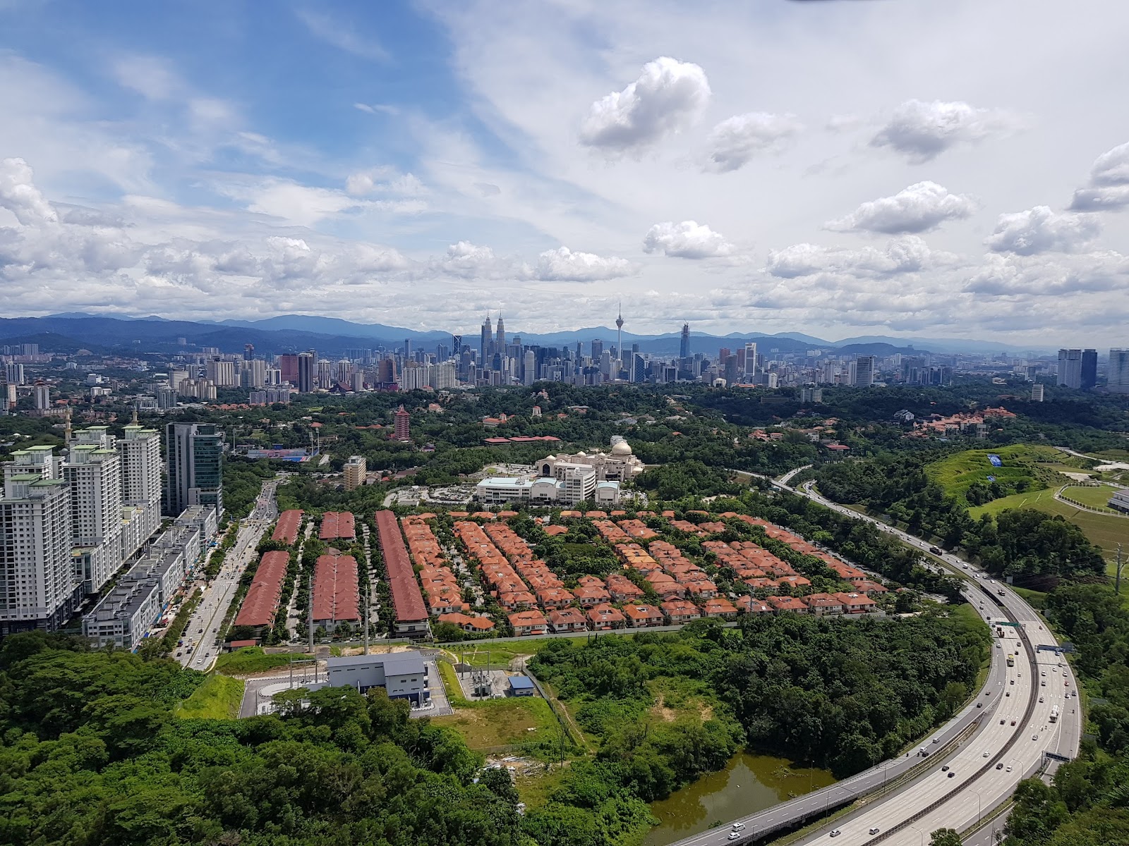 Seni Mont Kiara: Aerial view of Kuala Lumpur city skyline with KL Tower and Petronas Towers in the distance, foreground shows a residential township with red-roofed terrace houses, highway and greenery