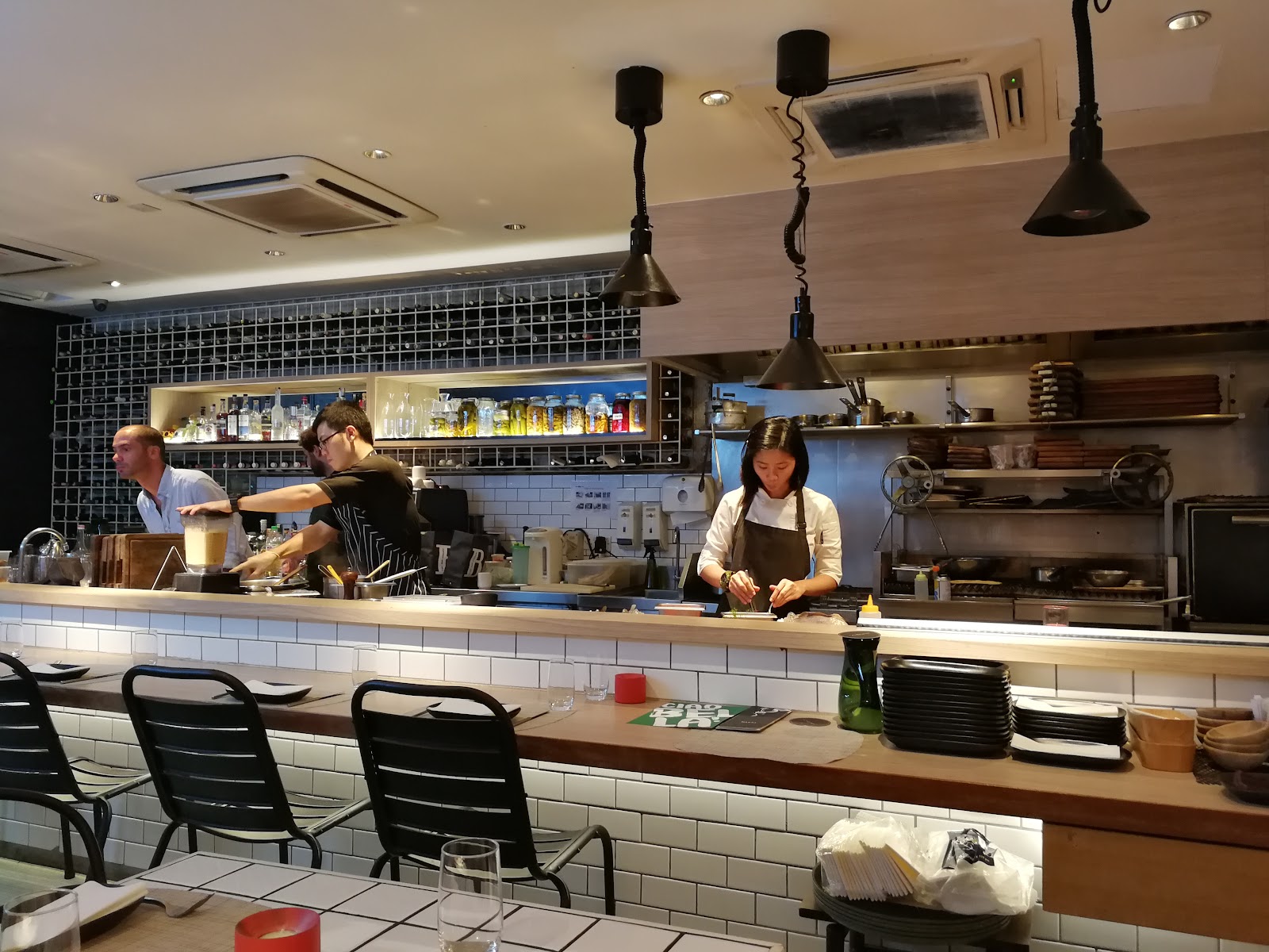 Pantai Towers: An open kitchen bar area with staff preparing food, bar seating, and shelves of bottles and jars in the background