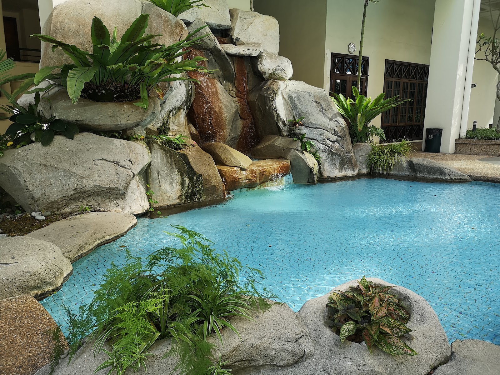 Araville: A resort-style swimming pool with a rock waterfall feature and tropical plants, next to a building with barred windows
